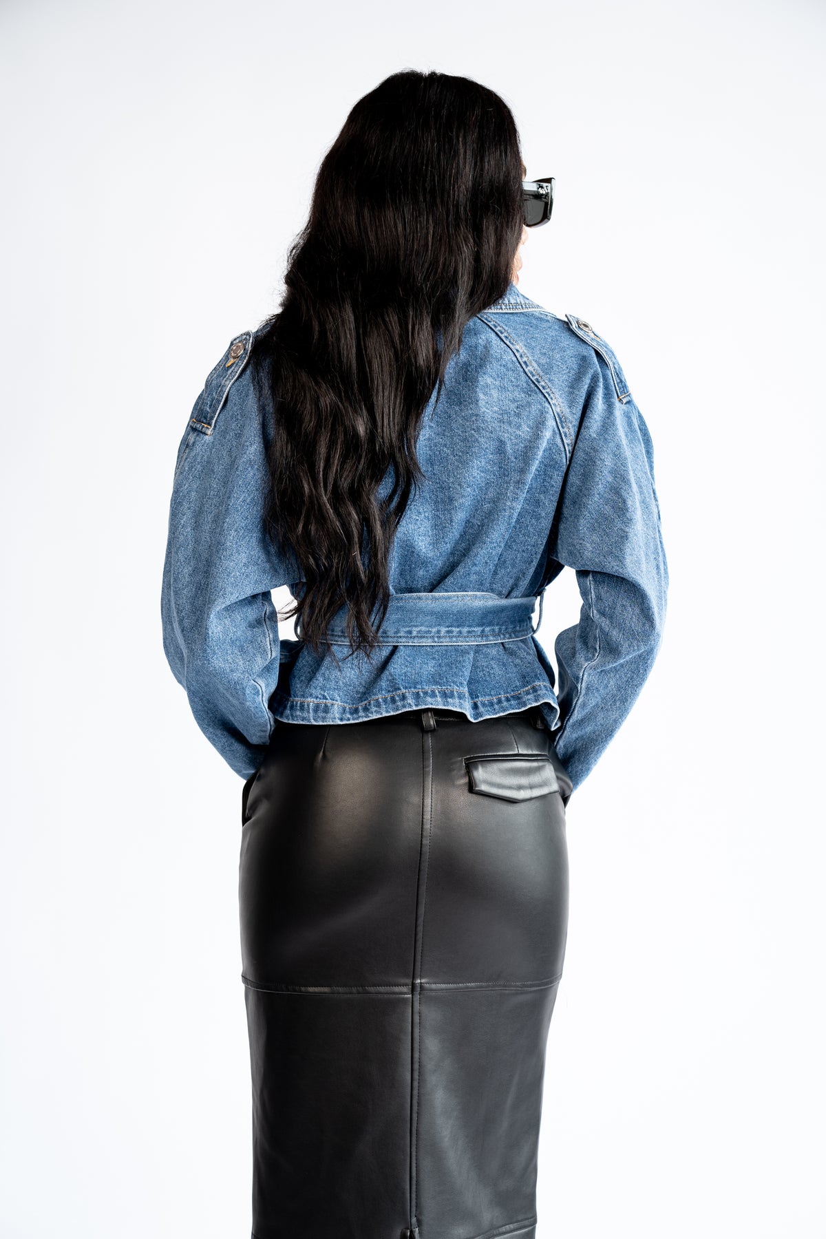 Back view of the blue cropped denim trench coat showing the shoulder epaulets, waist belt and flared hem.