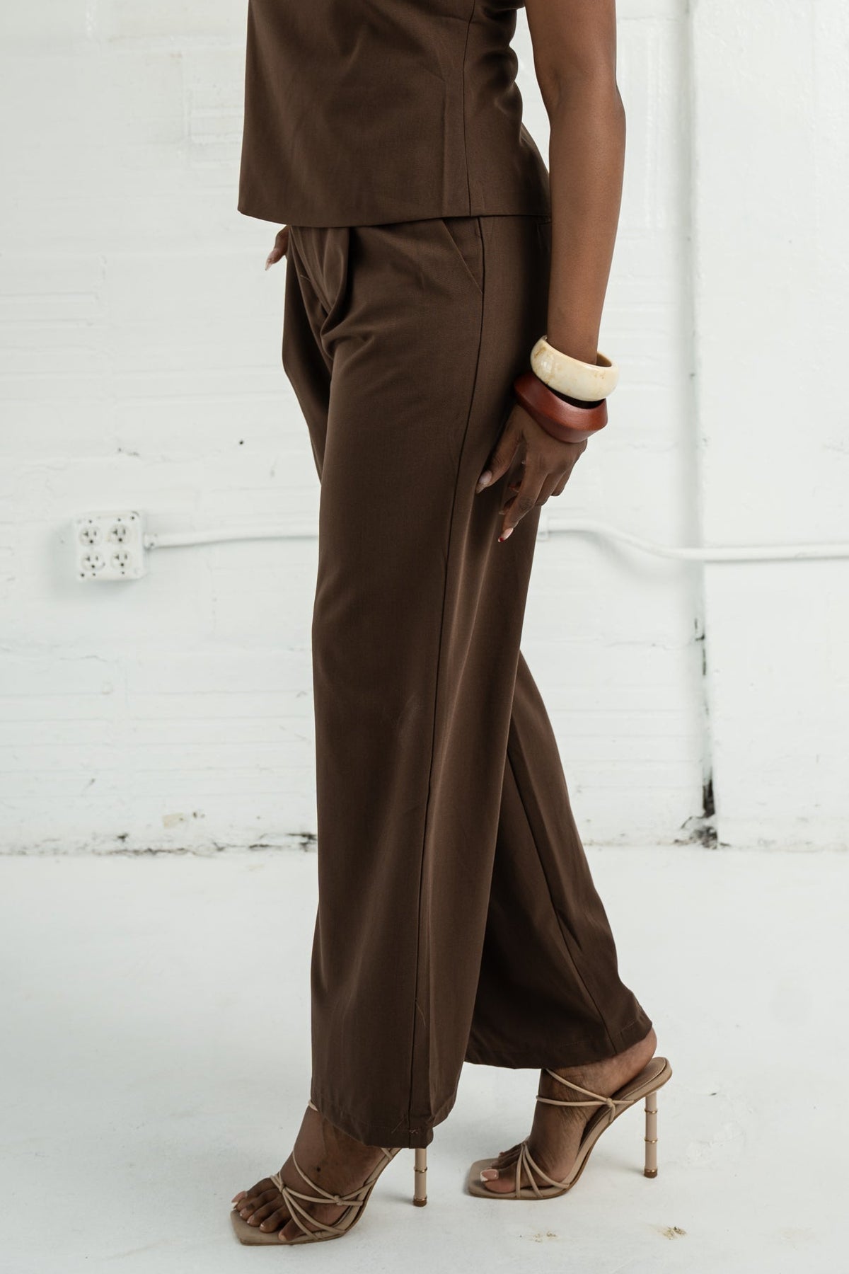 Woman wearing a brown sleeveless top and pants against a white wall.
