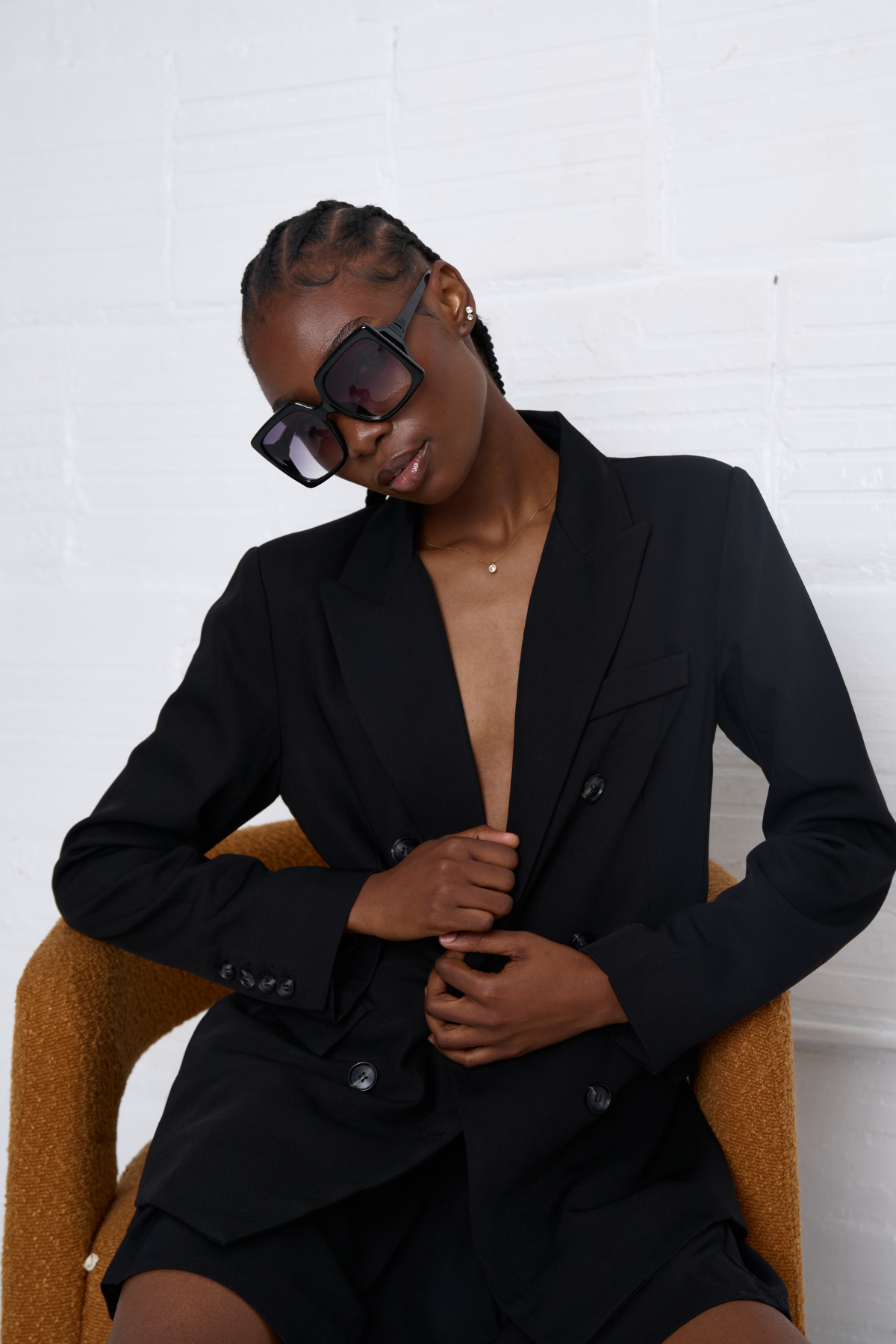 Woman wearing a black blazer and sunglasses sitting on a chair against a white wall.