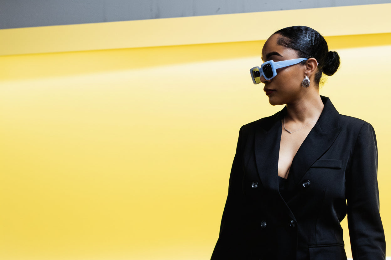 Woman wearing a black suit and blue sunglasses against a yellow and gray background
