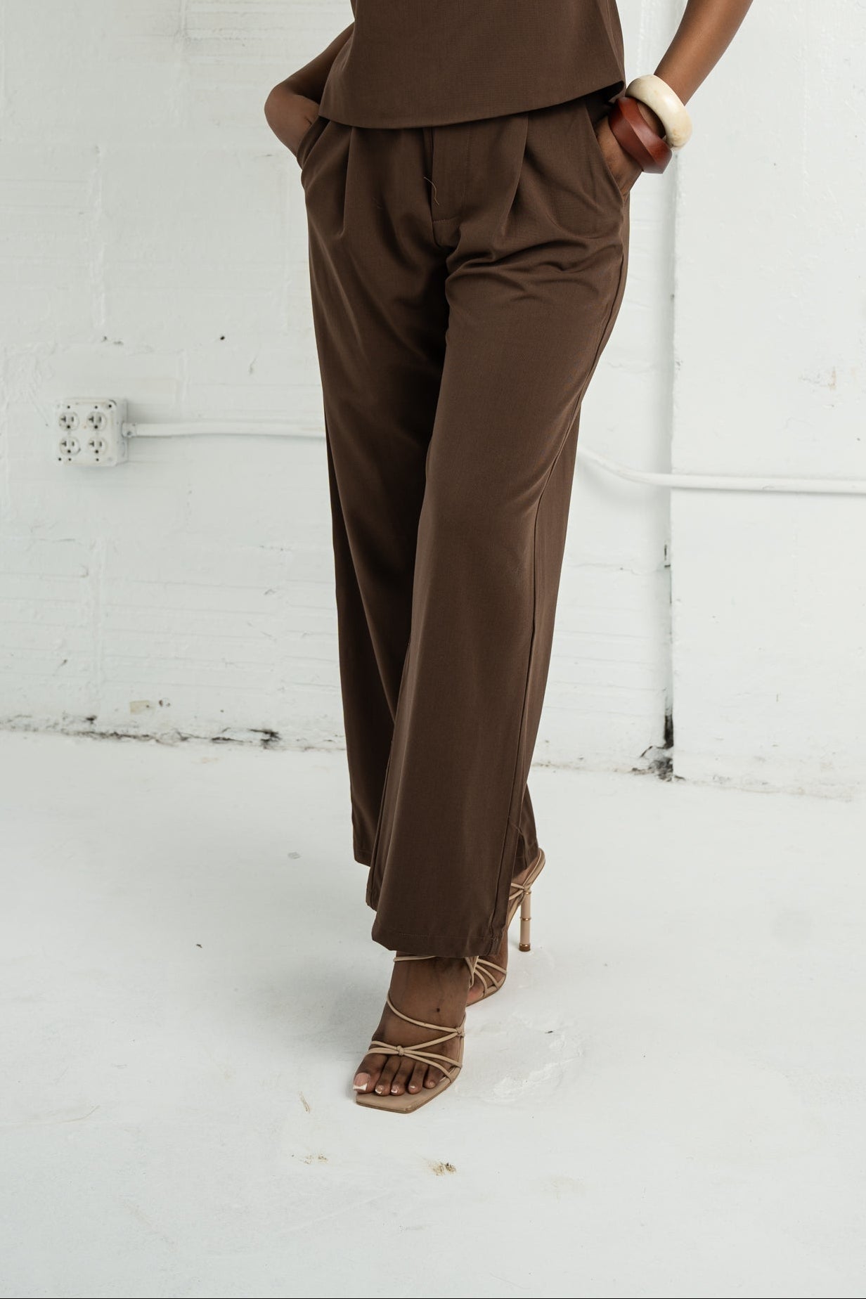 Woman wearing a brown sleeveless top and pants against a white wall.