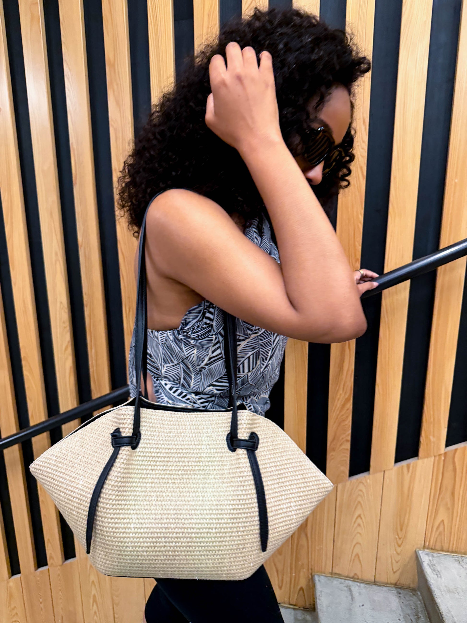 Woman holding a straw handbag with black straps against a striped wall.