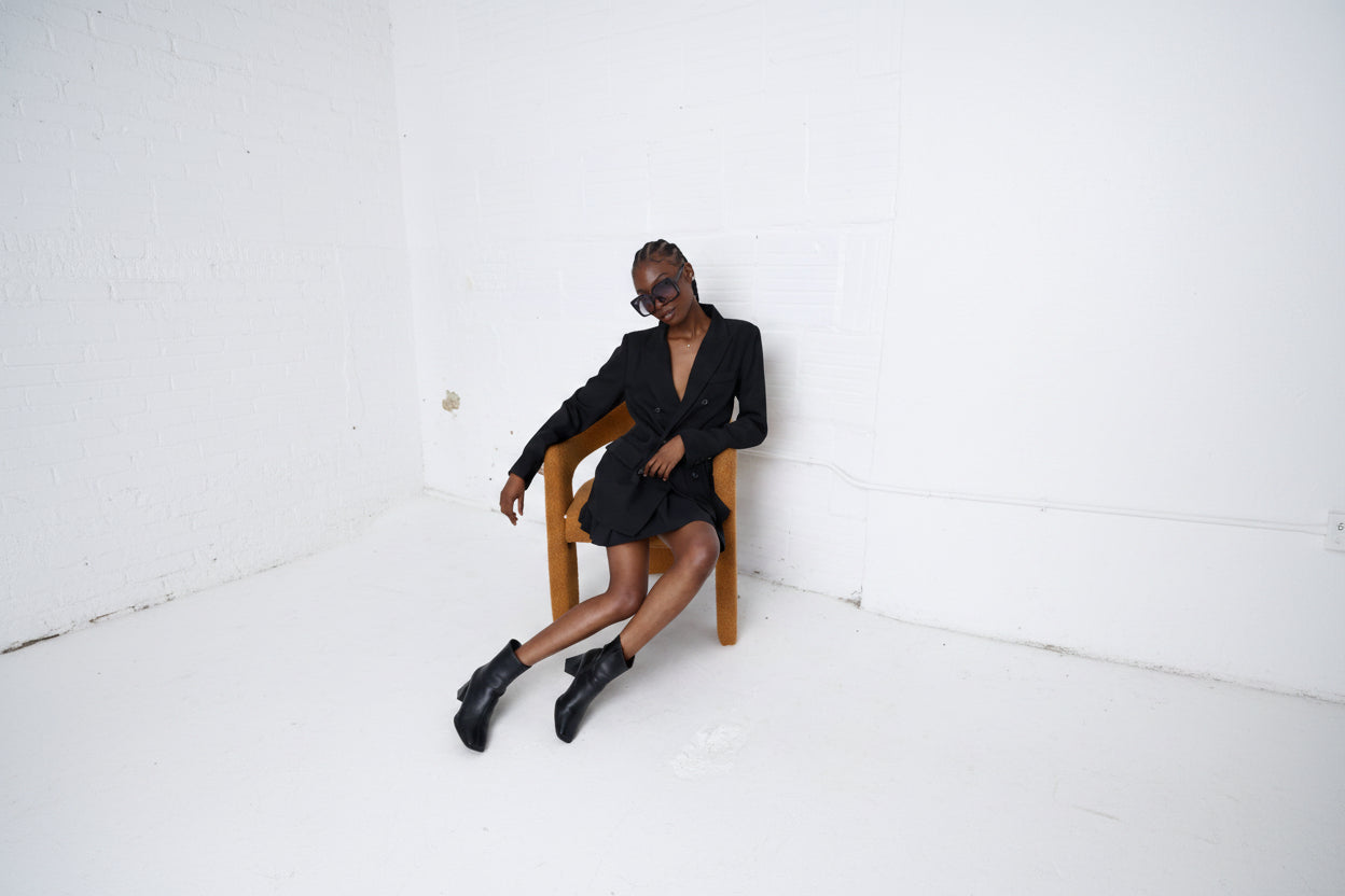 A model wearing a black double-breasted blazer dress and black leather ankle boots sits on a wooden chair against a white brick wall.