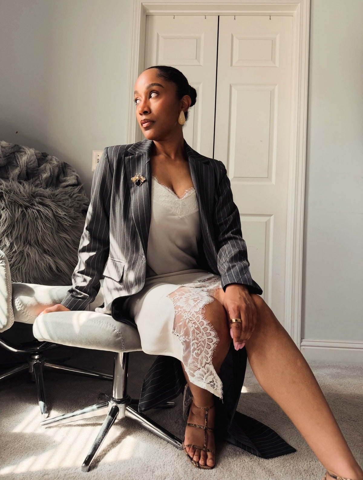 A woman sits on a white swivel chair in a room, wearing a gray pinstripe blazer over a white lace-trimmed camisole and white skirt. She is looking upward and to the side, with a white door behind her and a gray faux-fur chair visible to the left.